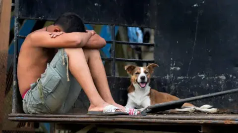 Getty Images A man taken into custody by border police agents during an operation against migrant smuggling waits on a truck next to a dog before being transferred near Santa Fe, Darien province, Panama, on August 7, 2024. 