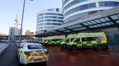 The front of a hospital building with a row of ambulances at the front and one police car. The sign above the door says Queen Elizabeth Hospital Birmingham