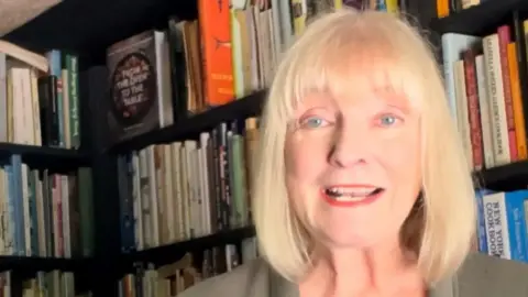 Ruth Watson A head and shoulders picture of Ruth Watson, who is sitting in front of a bookshelf with lots of books on it.
