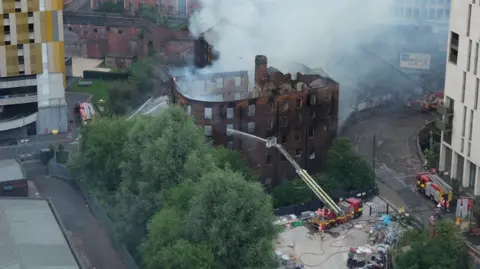 A drone aerial shot shows the smoking ruins of the Hotspur building in Manchester city centre. An aerial ladder platform can be seen spraying water into the building, which sits in the shadow of large tower blocks and next to a number of trees. 