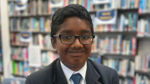 Sai has black short hair and wearing black rimmed glasses and is wearing a blue blazer, white shirt and blue and yellow tie. She is sitting in a classroom full of books.