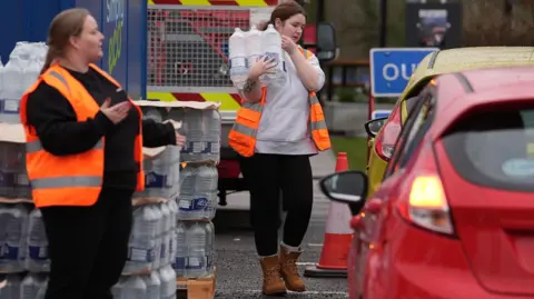 PA Media Two women wearing high‑visibility vests are working at an outdoor bottled water distribution point. One is carrying a multi‑pack of water towards a line of cars, while another stands near stacked pallets of bottles beside traffic cones and vehicles.