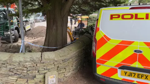 Malik Walton/BBC A police van parked next to a wall. Behind the wall a man can be seen operating a digger next to a mound of earth which looks to have been dug up.
