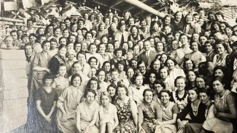 Saltaire World Heritage Education Association A black and white photo showing a large group of women dressed in everyday clothes. In the centre of the group stands a distinguished looking gentleman, in a suit and tie. 