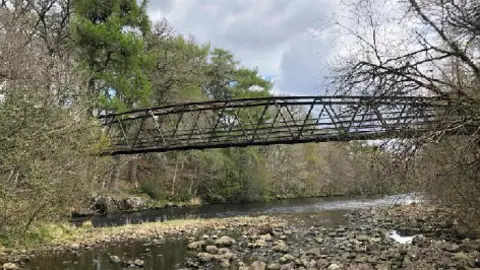 Forestry and Land Scotland A lattice metal footbridge a couple of metres high spans a river. Water levels are low and many rocks are visible. There is woodland on both banks of the river.