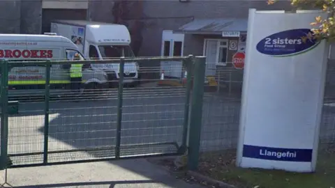 The outside of a factory, to the right is a sign that reads 2 sisters. To the left there is a green wired electric gate with a couple of vans parked inside.