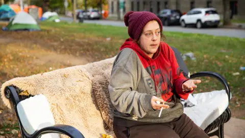 Samantha Nickerson sits on a park bench in downtown Halifax. She wears a burgundy toque, a grey and red hoodie and is speaking animatedly with a cigarette in hand 