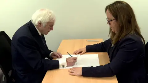 Jim and Dr Stephanie Rossit, who is wearing a black suit jacket, sat at a table. Jim is holding a pencil and is drawing