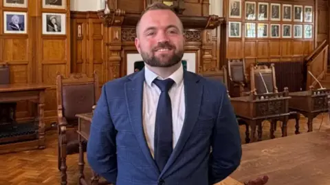 emma draper Devon stands, hands behind his back, inside a council chamber. He is wearing a navy suit and has short hair.