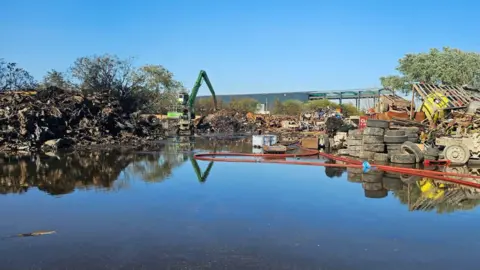 Nottinghamshire Fire and Rescue Service A scrap metal yard filled with piles of burnt metal which is surrounded by a large puddle of water. A fire hose can be seen on the right of the image.