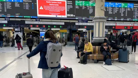 Woman standings looking at departure boards at train station holding two suitcases and wearing a backpack. On the board there's a big red sign that says: 'Radio System Fault Nationwide'