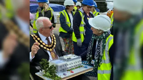 BSWICB Councillor Stephen Cooper wearing a black suit, black sunglasses and his mayoral chains. He is sitting down with a model replica of the treatment centre resting on his lap. He is surrounded by women wearing hard hats and yellow hi-vis vests on a construction site.