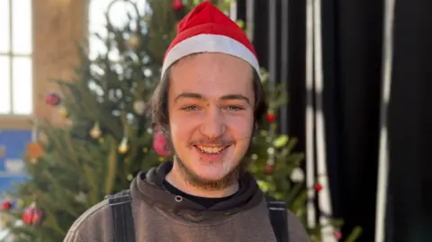 Charlie Jones/BBC A young man with facial piercings and wearing a santa hat smiles at the camera with a Christmas tree in the background