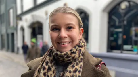 Lucy Kirgan is standing on Hill Street. She is wearing a brown coat and scarf. She has blonde hair. Over her shoulder is the red strap of a bag. Behind her are blurred buildings and people.