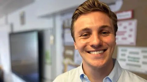 Jamie Wace is in a school classroom, standing and smiling in front a notice board. He has short light-brown hair and is wearing a light blue shirt with a white jumper over the top. There is a television on a wall behind him and some charts on a board, blurred in the background.