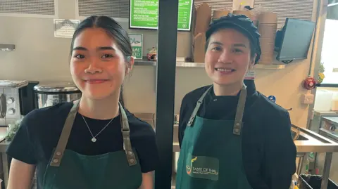 Two women smiling wearing aprons with kitchen behind them.