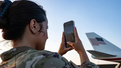 RAF Mildenhall A woman wearing a camouflage top is taking a photo on a mobile phone of a plane. The tail of the plane, with the stars and stripes flag on it, is in the background.