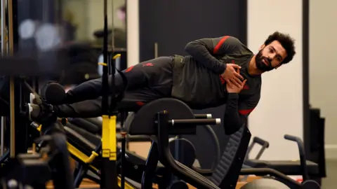Getty Images Mo Salah appears horizontal in a gym as he practices an exercise to strengthen his core. He is wearing a tracksuit and his hips are resting on a curved piece of gym equipment. He has his hands folded across his chess and a calm expression on his face.