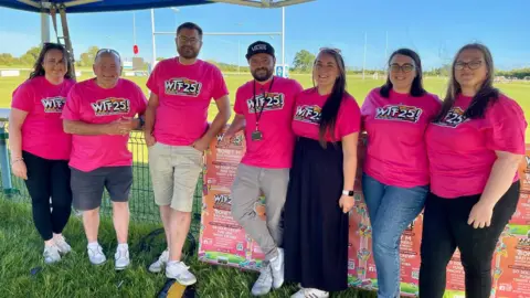Gary Cooke, second from left, and Nico Menghini, centre, co-organisers of The Wiltshire Throwback Festival with festival volunteers. They are standing in a field with bright pink festival t-shirts, smiling. 