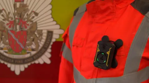 A firefighter in an orange high-vis jacket with a body-cam attached standing next to a logo for the Greater Manchester Fire and Rescue Service. 