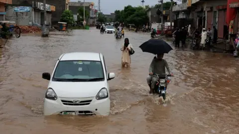रॉयटर्स ही एक कार, मोटारसायकल आणि लोक चालत आहेत - सर्व अंशतः पूर पाण्याने रस्त्यावर बुडलेले आहेत.