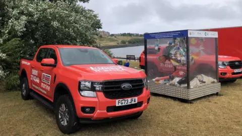 A red pickup is parked next to a sculpture made out of litter. Along the side of the vehicle it reads "Marine Emergency Response". A second red pickup is parked on the other side of the sculpture. In the distance the sea can be seen on the other side of a harbour.