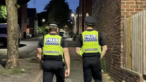 Two police officers walking through the Page Hall area of Sheffield. They are wearing high vis jackets and are walking on a road illuminated by streetlights.