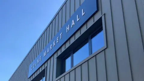 the outside of a building. It's modern looking, with overlapping grey vertical slates. Above two small windows a black sign reads "COLNE MARKET HALL"