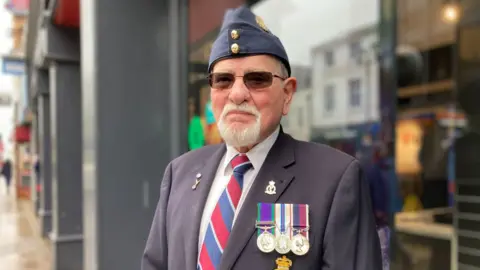 Shirley Henry / BBC George Dennis wears a navy hat and blazer with medals and military badges and red white and blue regimental tie. He has a white beard and is wearing tinted glasses.
