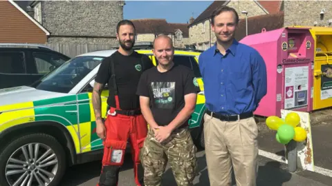 Francesca Harris Critical care practitioner Nick Richards, Mr Harris and councillor James Vitali stand next to each other posing for a photo and smiling at the camera. There is an ambulance 4 by 4 vehicle in the background and balloons to the right hand side to mark Mr Harris' start point. Mr Richards is wearing a black short sleeved t shirt with red cargo trousers, Mr Harris has a black t shirt with army camouflage trousers and Mr Vitali wears a blue buttoned shirt with a black belt and cream chino trousers
