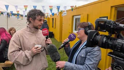 Tom Grennan in a light brown jacket andunglasses holding a red BBC CWR microphone and a cup. He is speaking to a female presenter, with a TV camera in the foreground on hte right. There are yellow demountables and white marquees in the background