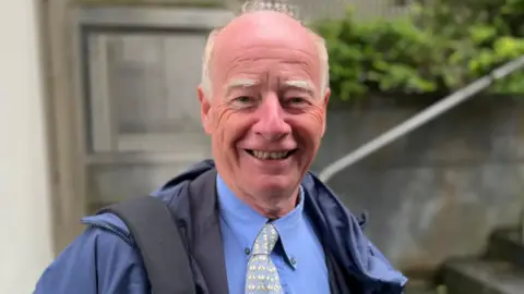 A man wearing a blue shirt with a tie and a dark blue jacket on top, smiling at the camera. The background of stairs and a door is blurred.