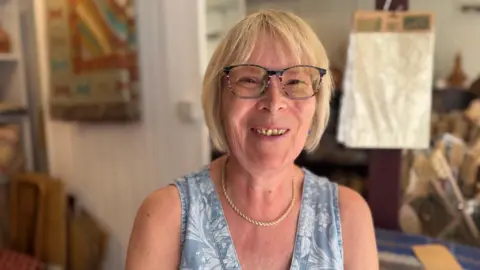 Liz wears glasses and has her blond hair trimmed in a bob. She wears a blue and white top with gold necklace and smiles for the camera. Material and soft furnishings are blurred in the background.