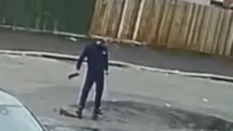Greater Manchester Police A grainy screenshot from CCTV footage of a man in a dark tracksuit standing in a puddle in the street.