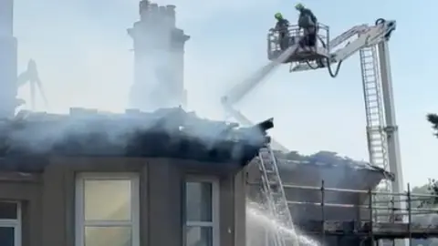Firefighters on an aerial platform above a smouldering house.