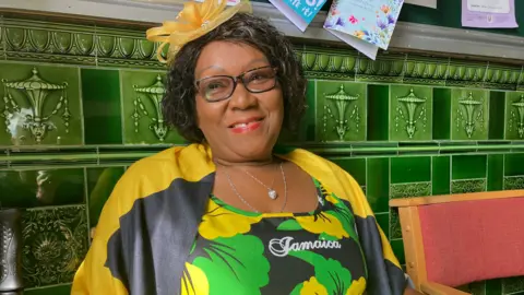 BBC/Phil Bodmer A woman in a colourful floral top with the word "Jamaica" printed above her chest in silver. She is wearing glasses and two silver necklaces as she sits in front of a wall fitted with green tiles.