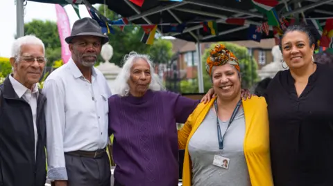 Two men and three women, including Shirley Williams, pose for a photograph at a Windrush event from 2024. They stand under a gazebo draped in flags from a variety of nations.