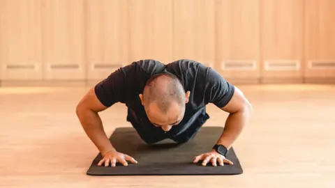 A man is doing push up on the yoga mat. He is in a room with a wooden floor and walls behind him.