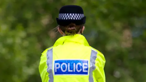 A female police officer, with her hair tied back in a low bun and wearing a hat, is seen from behind. She is wearing police uniform with the word POLICE written on the back