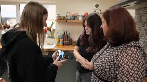 Sue, a middle-aged woman with dark red hair, stands in her kitchen with two of her daughters. They are looking at a mobile phone.