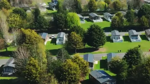 An aerial view of Allerthorpe holiday park featuring multiple cabins arranged in neat rows. Each unit is surrounded by lush green lawns, with a mix of evergreen and deciduous trees scattered throughout the area. Paved pathways connect the houses, and some units have vehicles parked beside them. The grass appears freshly trimmed.