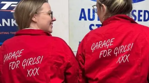 Hannah Simpson Two women stand with their backs to the camera showing their red overalls with 'Garage for Girls! xxx' written on the back