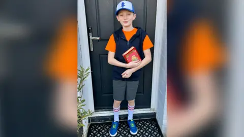 Freddie is wearing an orange T-shirt and black, sleeveless hoodie. He is also wearing grey shorts and long, white shorts with red banding and blue trainers. Freddie is standing on a doorstep of a house.