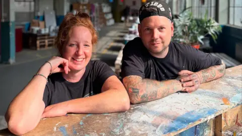 Seagulls Cat Hyde and Steve Dennis, a woman with ginger hair tied in a bun and a man, both in black t shirts splatered with paint, the man is also wearing a cap. They are in a large open plan warehouse which sells paint.