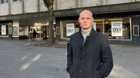 Max Wilkinson wears a button up coat and roll neck sweater outside a closing department store in Cheltenham