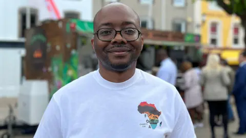 Washington is wearing glasses and a white T-shirt with a colorful Africa graphic stands outdoors in a town square. Trees and pastel-colored buildings are visible in the background, along with a small food stall and several people gathered behind.