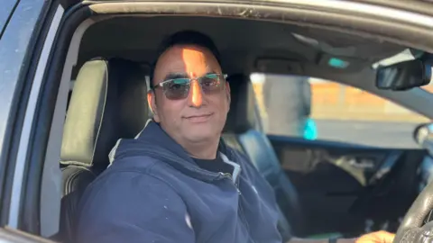 A man in a taxi. He is wearing a blue hoodie and has sunglasses on.