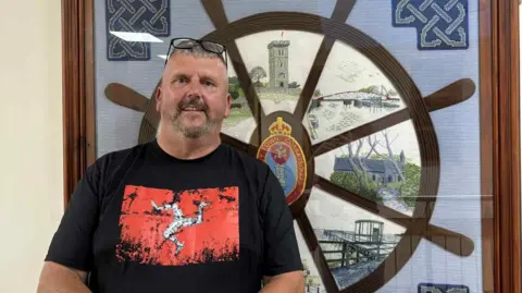 Chris Martin, a man with grey hair who has a beard and moustache, smiles with reading glasses on his head. He is wearing a black t-shirt with the red-and-white Manx flag in the middle.