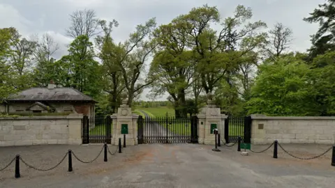 Google Maps The gated entrance to the hotel in Ballyfin Demense. There is a stone wall with black iron gates, a gatehouse and a long path through a large lawn.