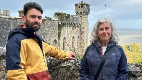 Sky Benjamin and Sandra Preiss standing against a wall with Gwyrch Castle in the background. Benjamin wears a blue, white and red thick stripe coat. Sandra wears a dark blue coat. 